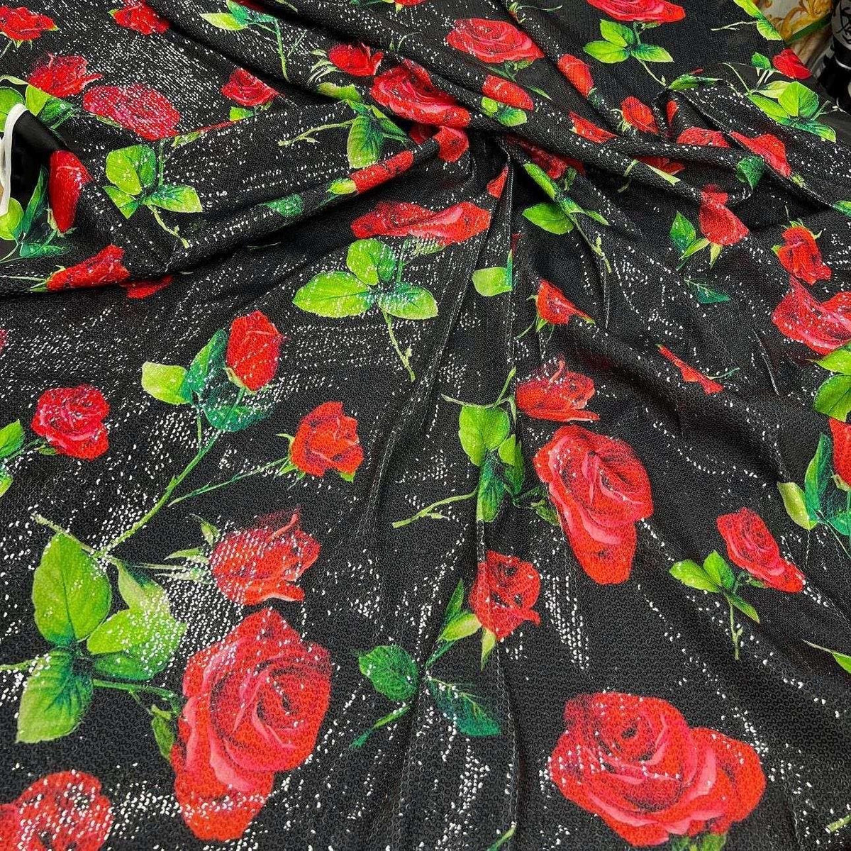 Black fabric with red rose pattern and green leaves, covered in shimmering sequins with two-way stretch, ideal for elegant evening wear.