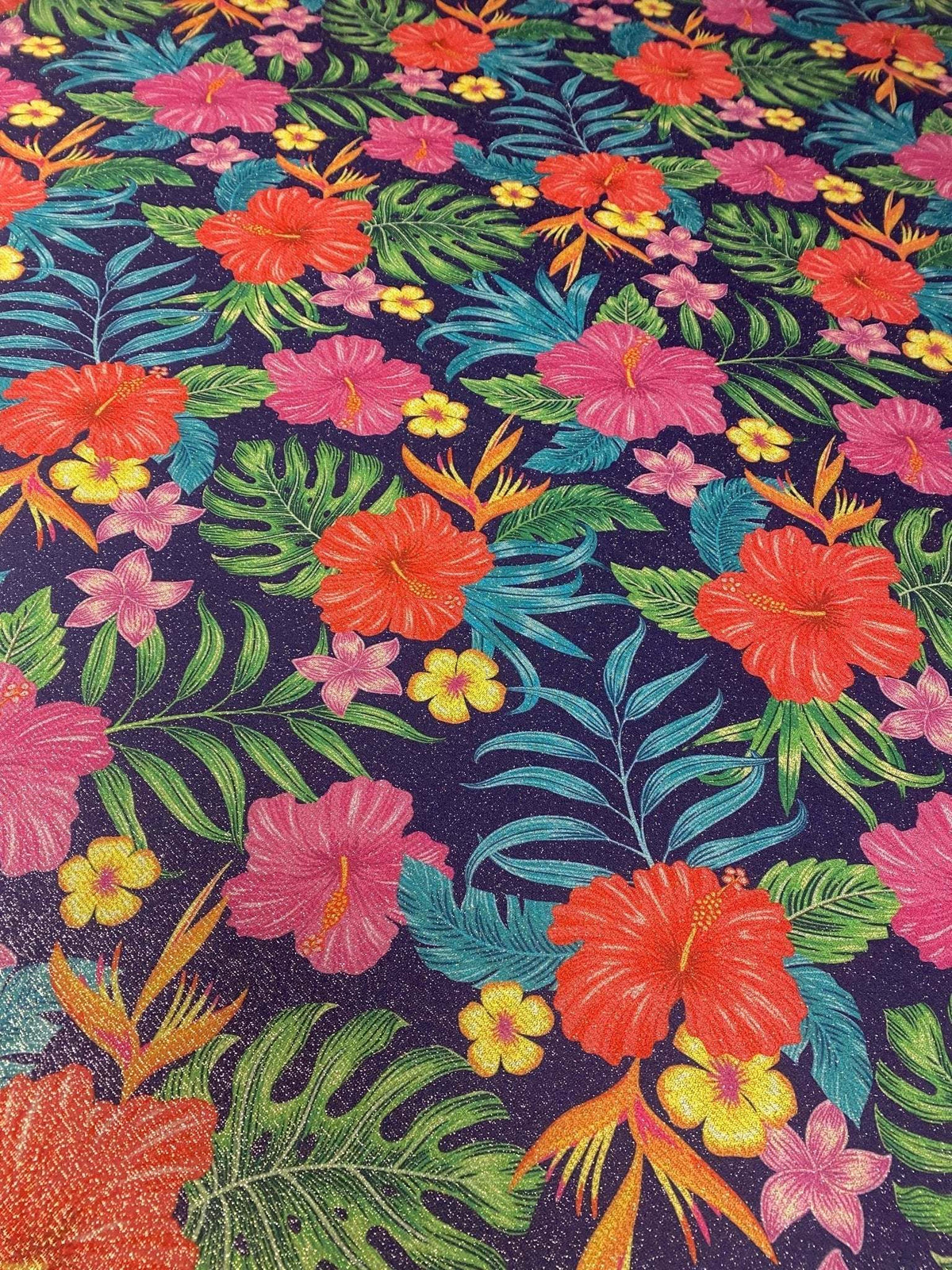 Tropical floral fabric with vibrant hibiscus and tropical leaves pattern on dark background, featuring gold lurex striped glitter accents and 4-way stretch polyester blend.